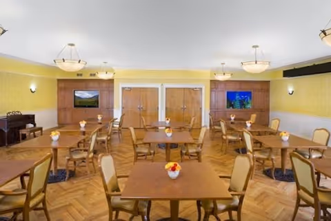 Bright dining room with multiple wooden tables and chairs on a parquet floor, chandeliers overhead, and double doors at the far wall.