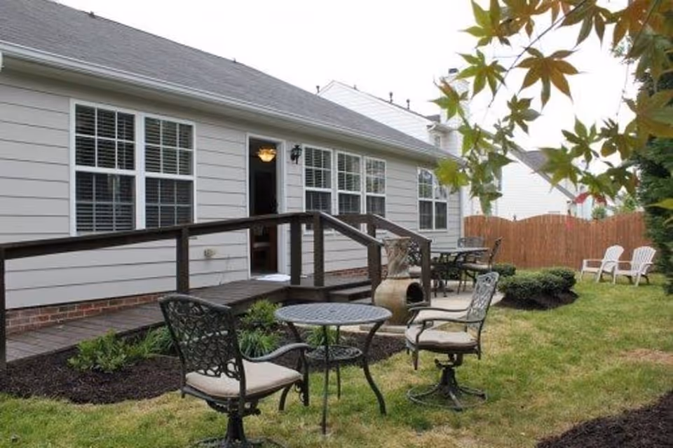 Backyard patio with metal table and chairs, a wooden ramp leading to the house, and a grassy yard with additional seating.
