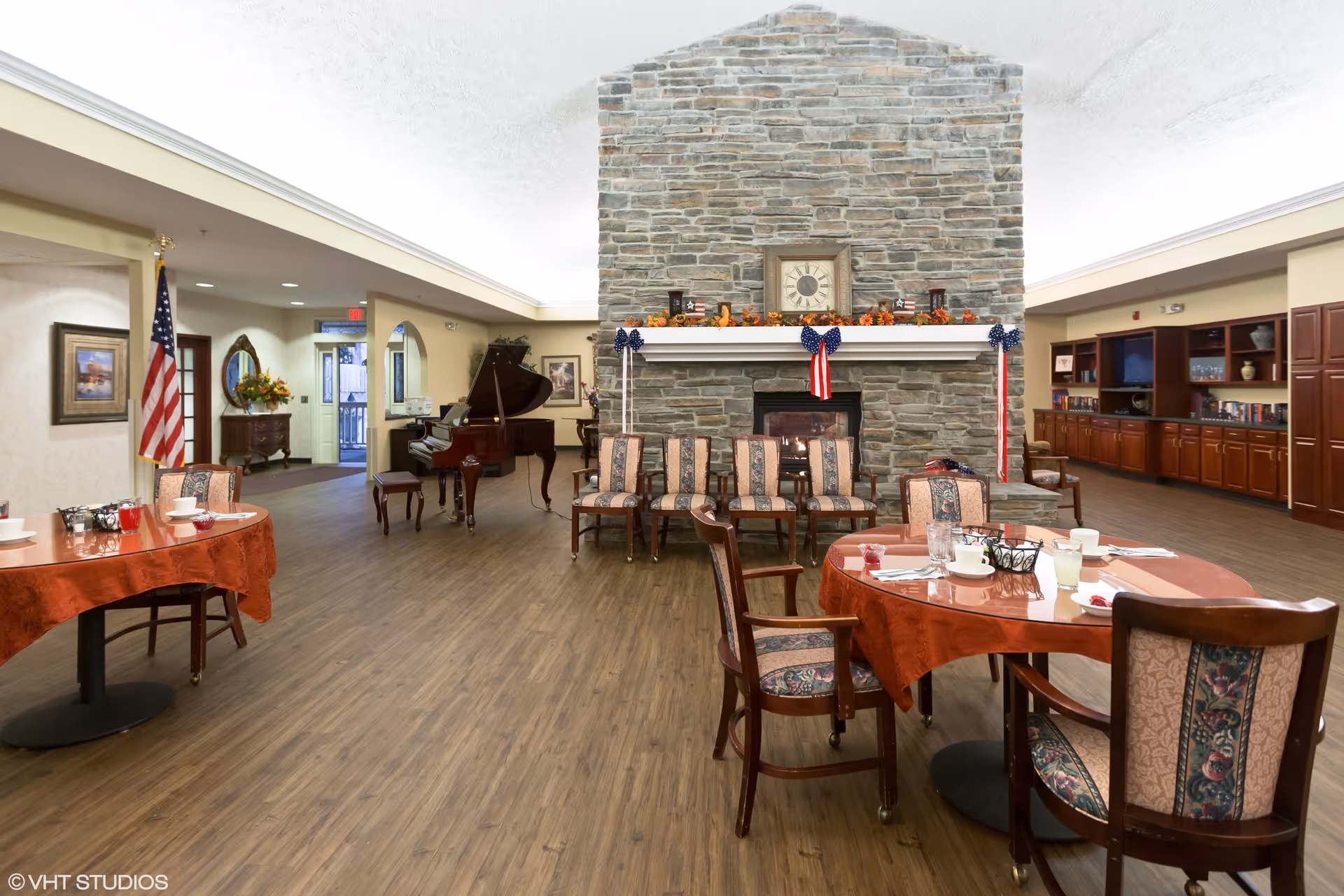 A spacious common area with a large stone fireplace decorated with patriotic ribbons and a clock on the mantel. Several chairs are arranged in front of the fireplace. To the left, there is a grand piano and an American flag. Two round tables with red tablecloths and chairs are set with cups and glasses. The room has wood flooring and built-in wooden cabinets along the right wall.