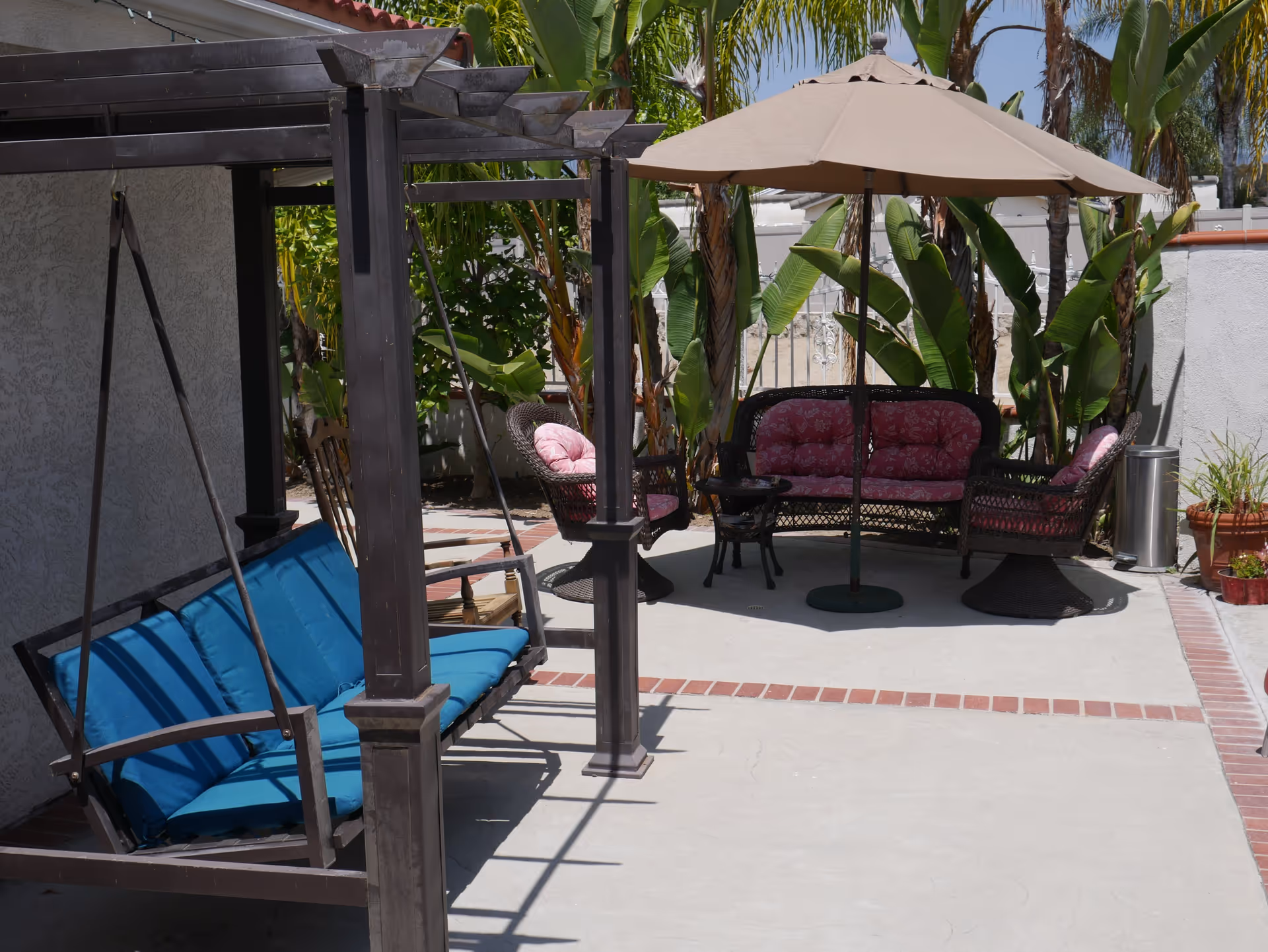 Outdoor patio area with a wooden swing bench with blue cushions on the left, and a seating arrangement with a wicker loveseat and two chairs with pink cushions under a large beige umbrella. There are tropical plants and palm trees in the background, and the area is paved with concrete and bordered with bricks.