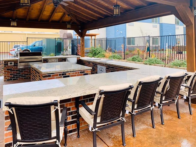 Outdoor covered patio area with a built-in brick and granite countertop kitchen and bar seating. Six cushioned chairs are lined up along the counter. In the background, there is a black metal fence, some greenery, and a blue building.