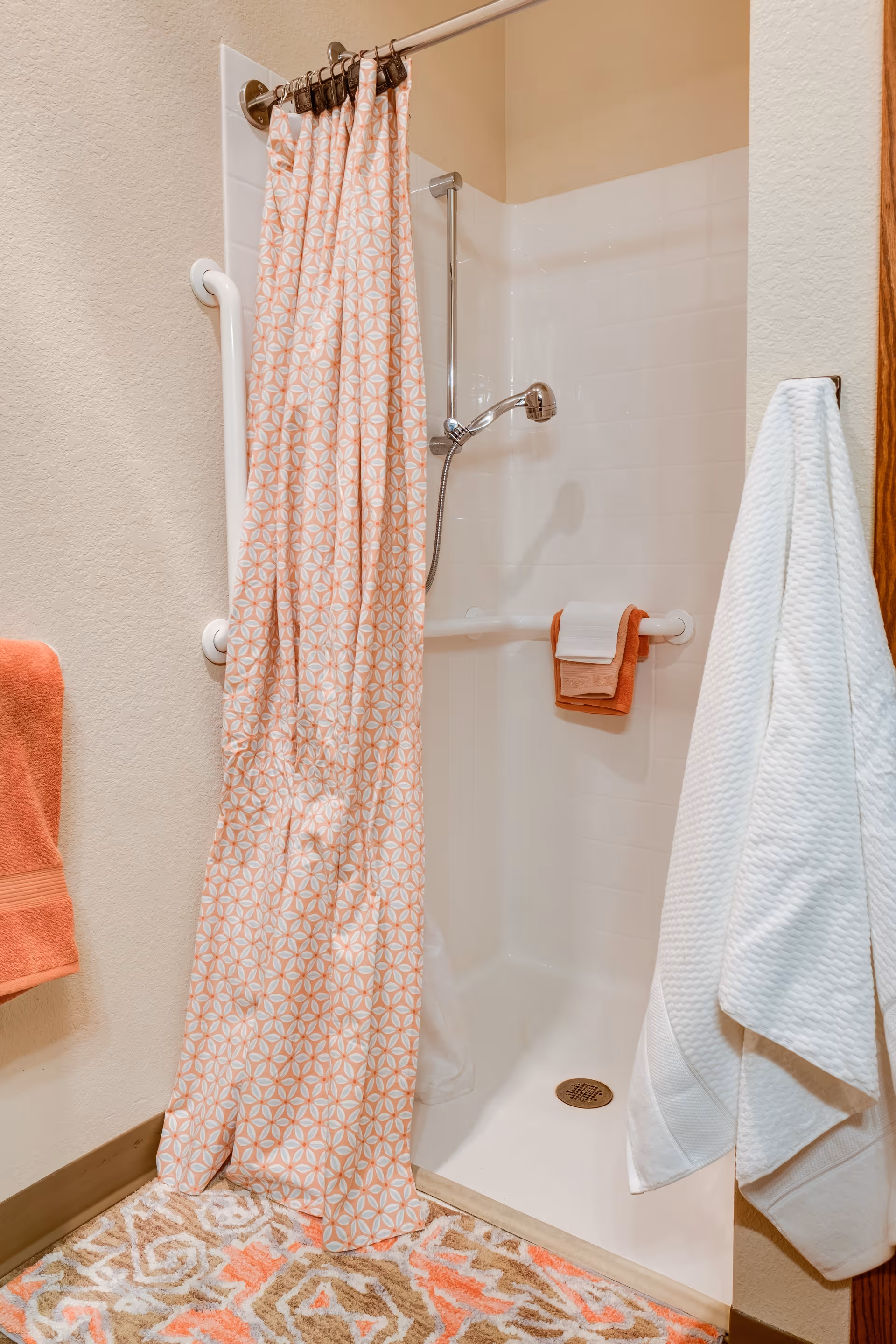 A small shower area with white tiled walls and a handheld showerhead. There is a white grab bar on the wall and a peach and white patterned shower curtain partially drawn. Two towels, one white and one peach, hang on the grab bar inside the shower. Outside the shower, a white towel hangs on a hook and a peach towel is draped over a rail. The floor has a patterned rug in shades of peach, beige, and white.