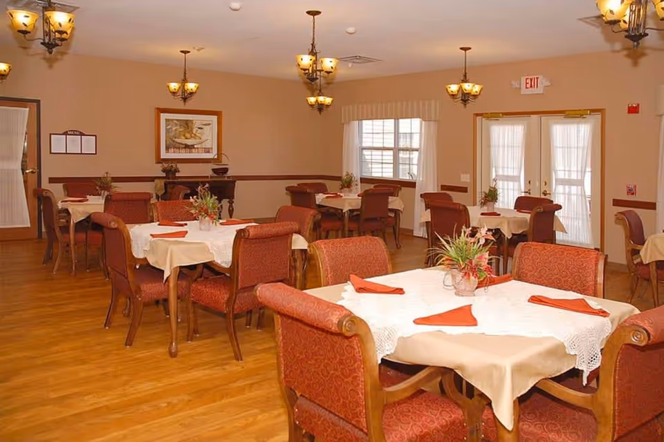 A dining room in a senior living facility with multiple tables covered with beige tablecloths and white lace overlays. Each table has red cloth napkins and a floral centerpiece. The room has wooden flooring, beige walls, several hanging light fixtures, and windows with white curtains. There are double doors with glass panels and an exit sign above them.