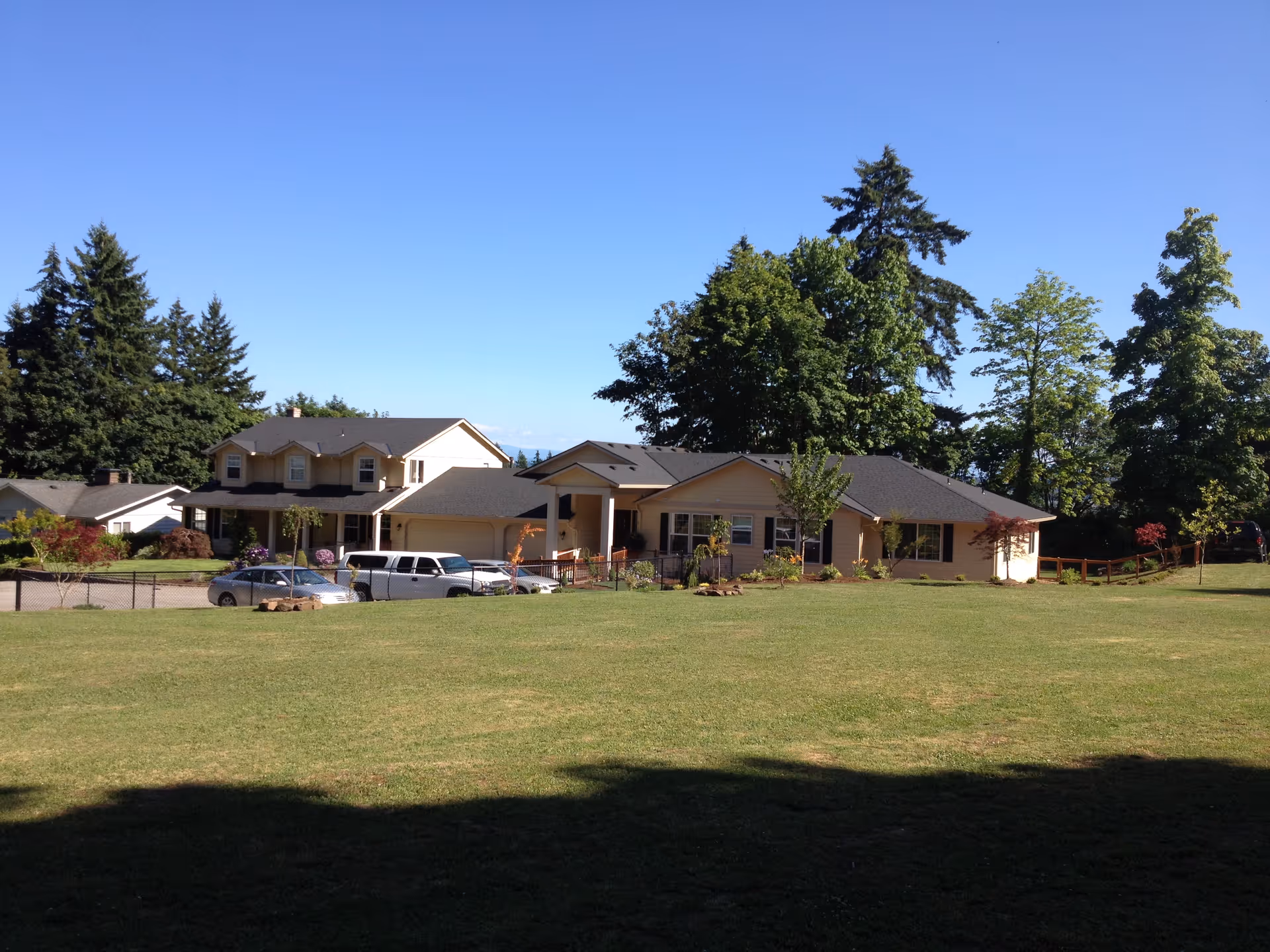 A large, single-story residential building with a dark roof and beige exterior, surrounded by a well-maintained lawn and several trees under a clear blue sky. There are a few parked cars in front of the building, and a two-story house is visible to the left.