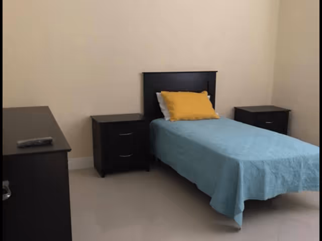 A simple bedroom with a single bed dressed in a blue bedspread and yellow pillow, flanked by two dark nightstands and a dresser.