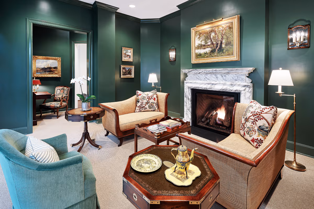 A cozy living room with dark green walls, a white marble fireplace with a fire burning, two beige sofas with floral cushions, a teal armchair, and wooden tables with decorative items including a teapot and a plate. The room is softly lit by wall sconces and floor lamps, and there are framed paintings on the walls.