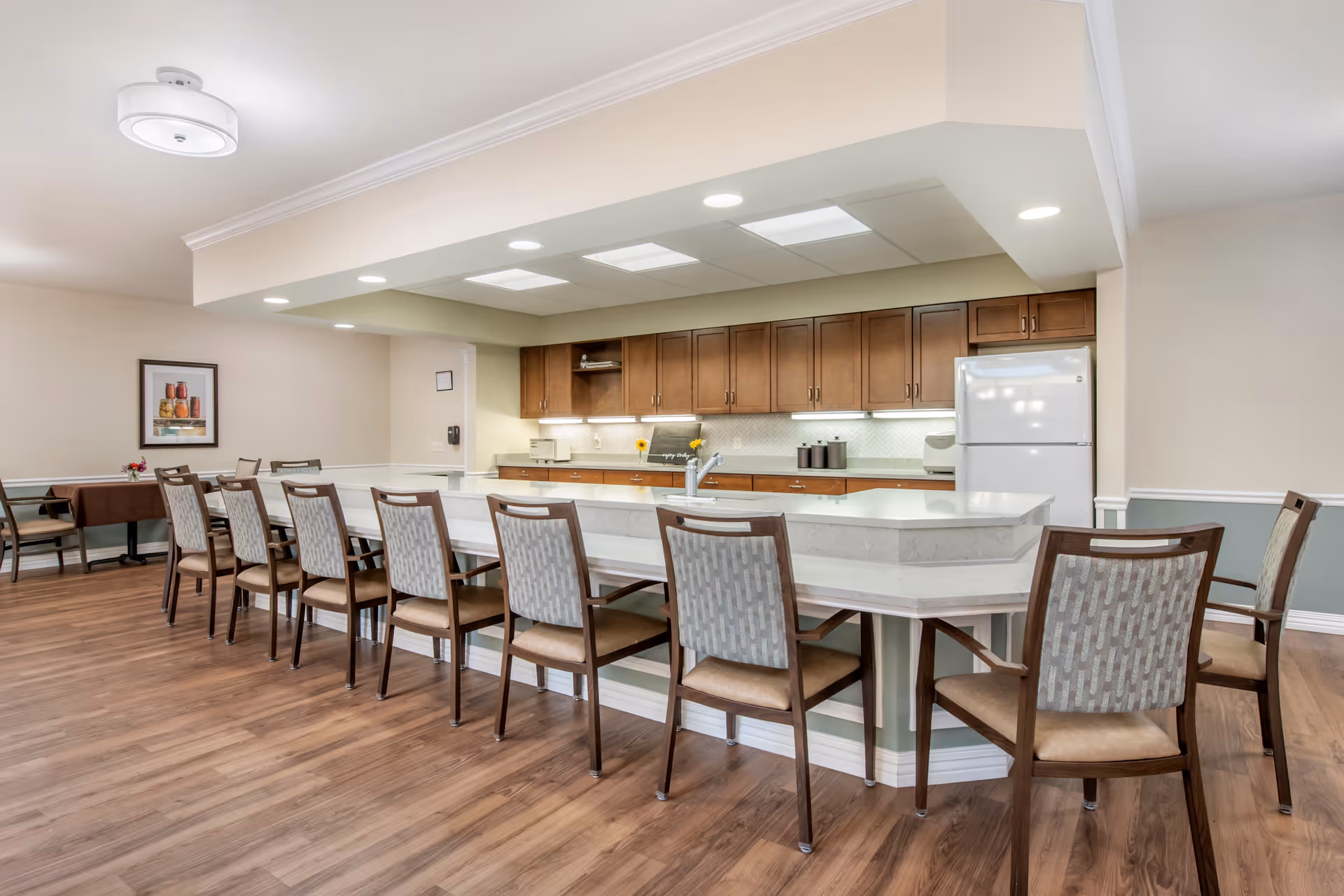 A spacious kitchen and dining area with a long white countertop island surrounded by wooden chairs with patterned upholstery. The kitchen features wooden cabinets, a white refrigerator, and under-cabinet lighting. The room has wood flooring, recessed ceiling lights, and a framed picture on the wall.