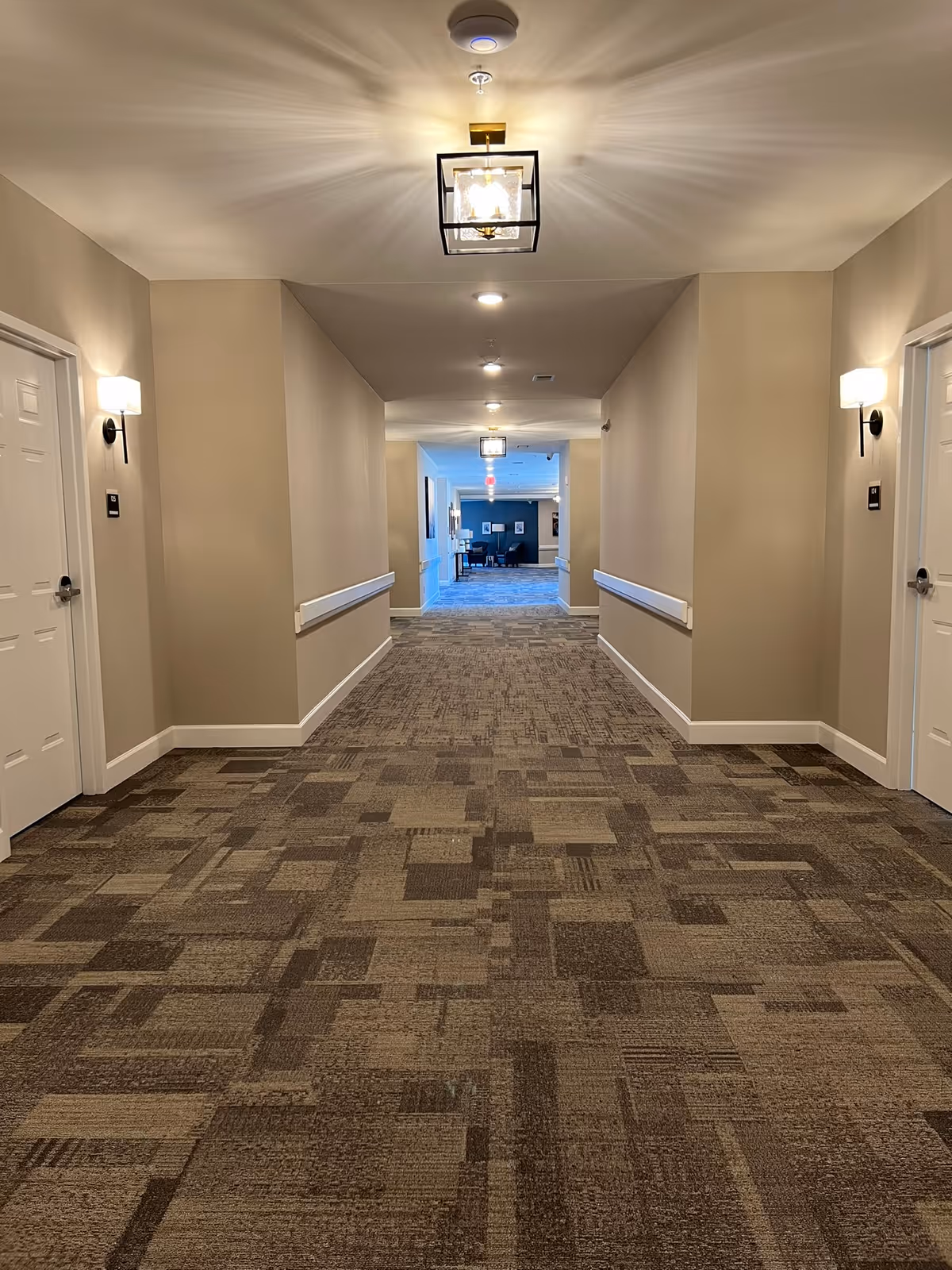 A well-lit hallway in an assisted living facility with beige walls, patterned carpet flooring, white doors on both sides, wall-mounted lights, and ceiling light fixtures. The hallway leads to a common area with seating visible in the distance.