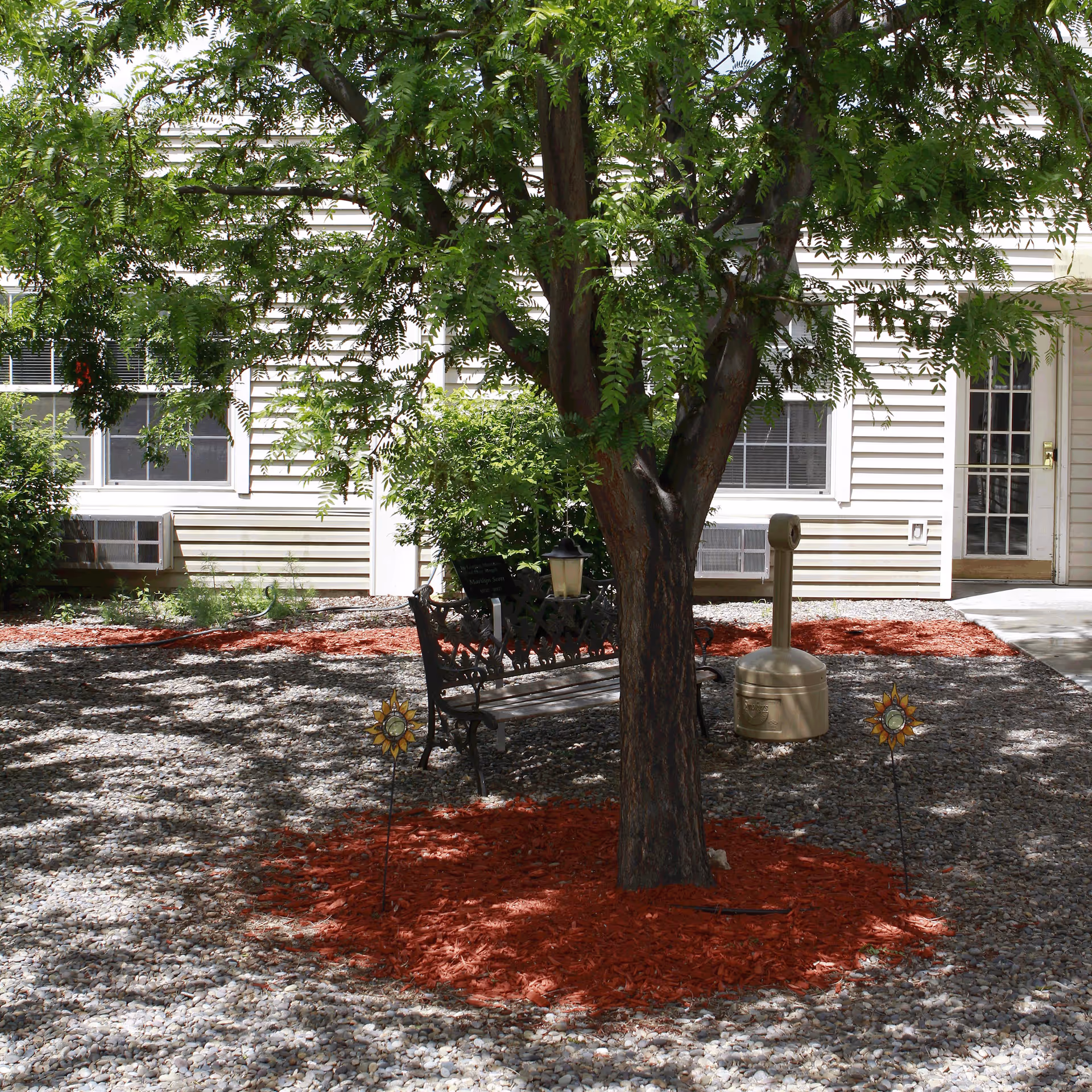 Outdoor garden area with a large tree surrounded by red mulch and gravel. There is a black metal bench with a small lantern on it, two decorative sunflower stakes, and a beige outdoor ashtray. The background shows a white building with windows and a door.
