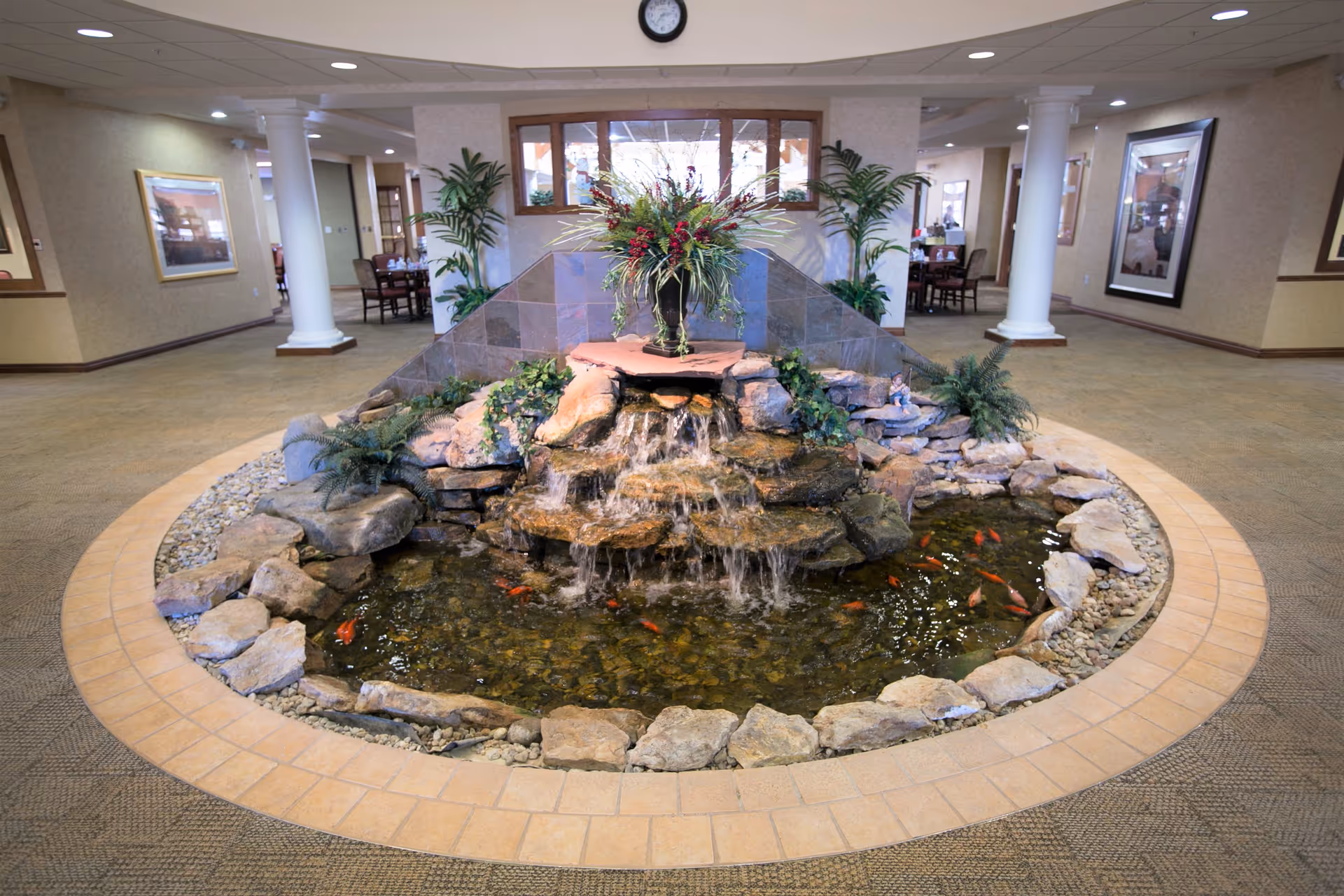 Indoor water feature with a small waterfall flowing over rocks into a pond with orange fish, surrounded by plants and decorative stones, located in a spacious hallway with carpeted floors, columns, framed artwork on the walls, and seating areas in the background.