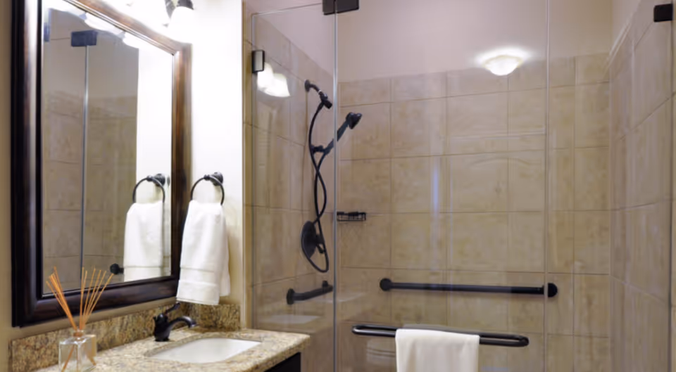 A bathroom with beige tiled walls and a glass-enclosed shower. The shower has a black showerhead and grab bars. There is a granite countertop with a sink, a dark-framed mirror above it, and a towel hanging on a ring next to the mirror. A reed diffuser is placed on the countertop.
