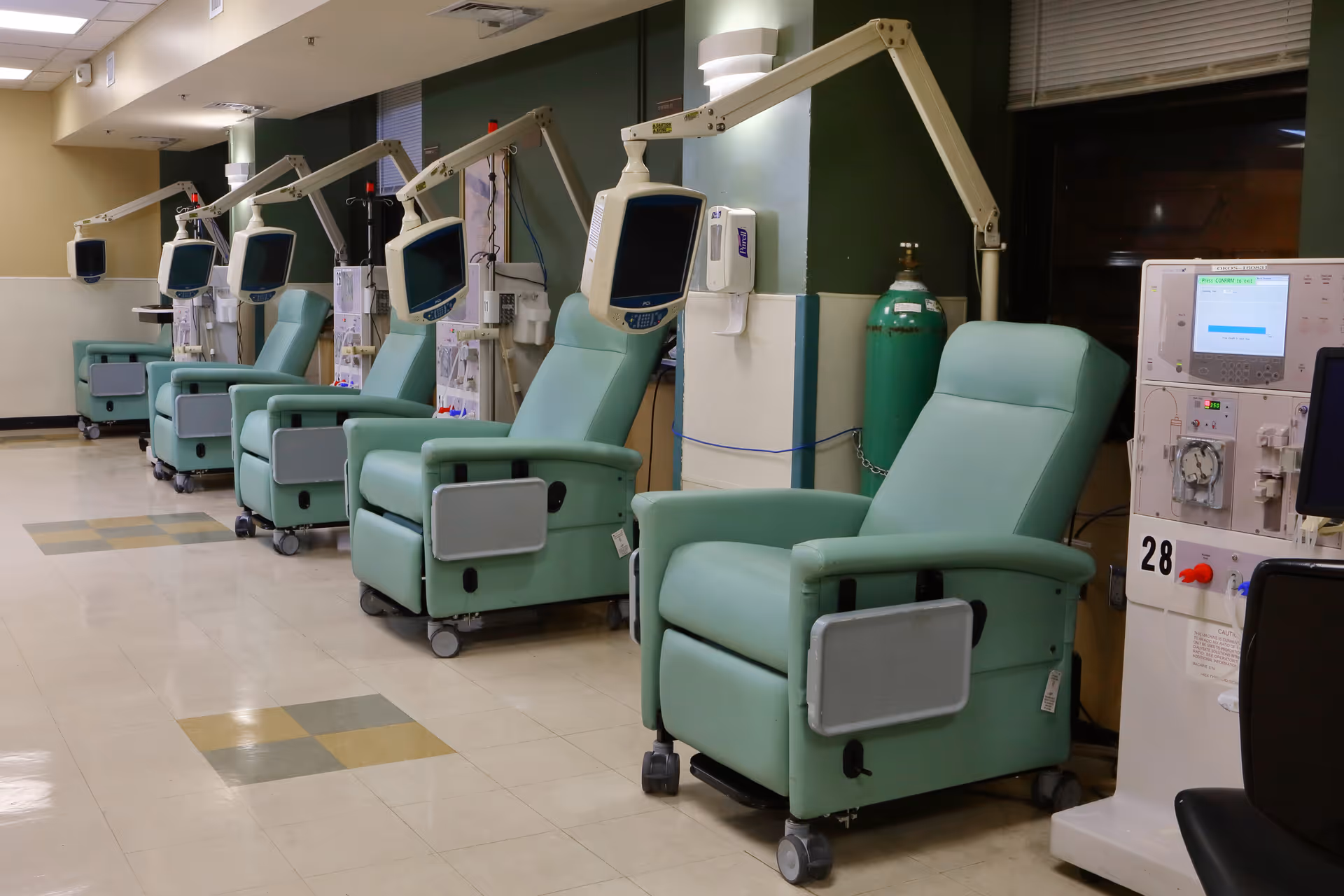 A row of green reclining medical chairs with attached monitors and medical equipment in a clinical setting, likely used for dialysis or similar treatments. The room has tiled flooring and medical gas tanks are visible against the wall.