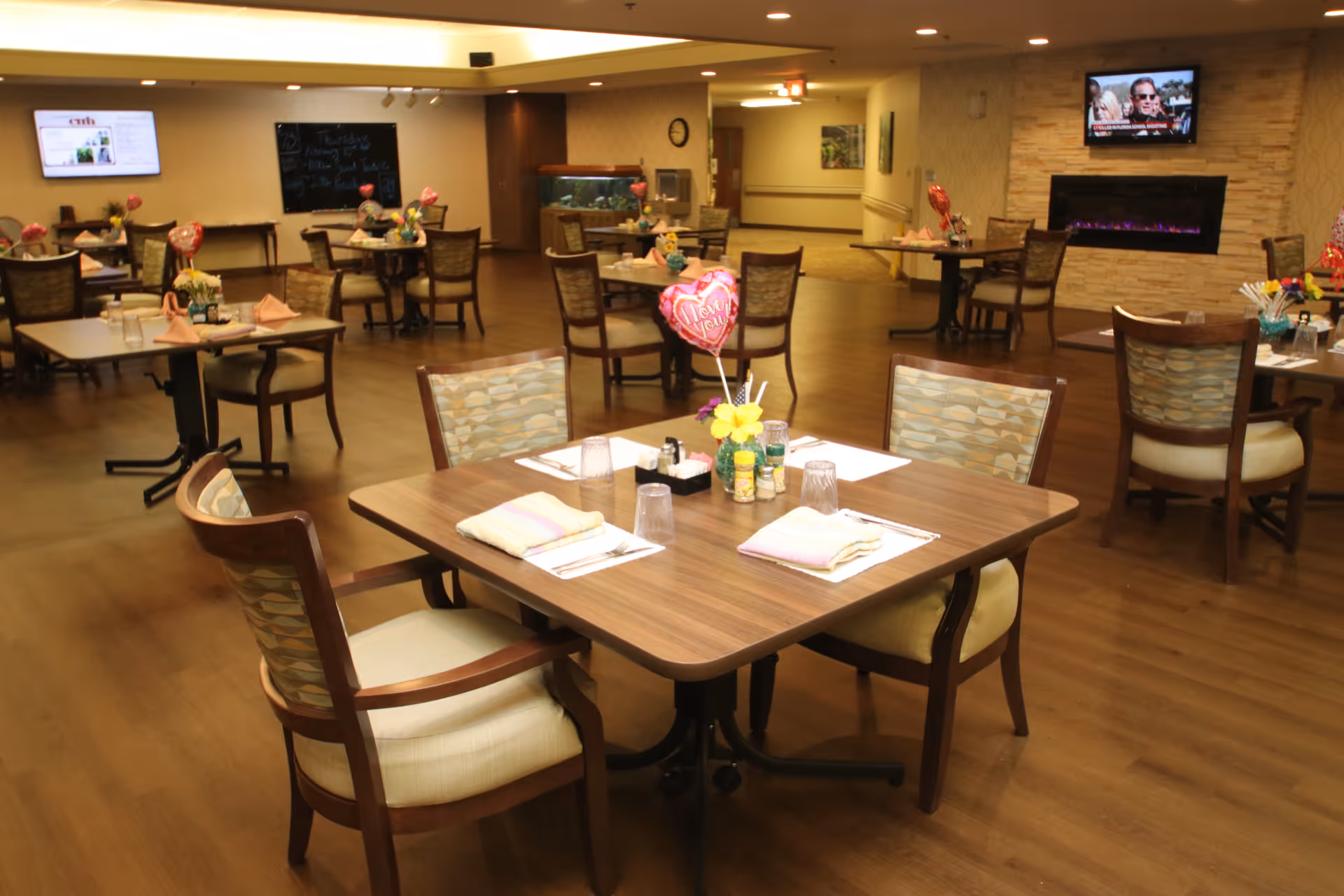 Spacious dining room with set tables and chairs, floral centerpieces and balloons, a wall-mounted TV and fireplace in the background.