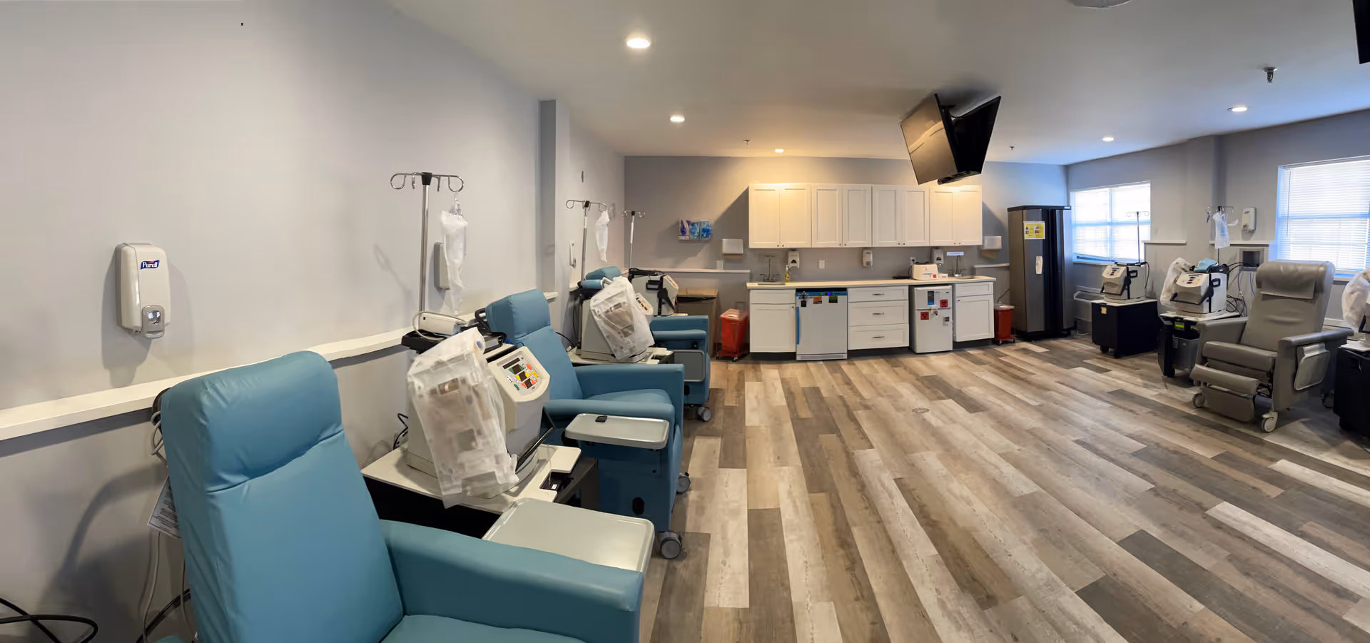 A spacious medical treatment room with multiple blue reclining chairs arranged along the walls, each equipped with dialysis machines and IV stands. The room has light-colored walls and wood-patterned flooring. There are white cabinets, a refrigerator, and a wall-mounted TV in the background. The room is well-lit with ceiling lights and natural light from windows.