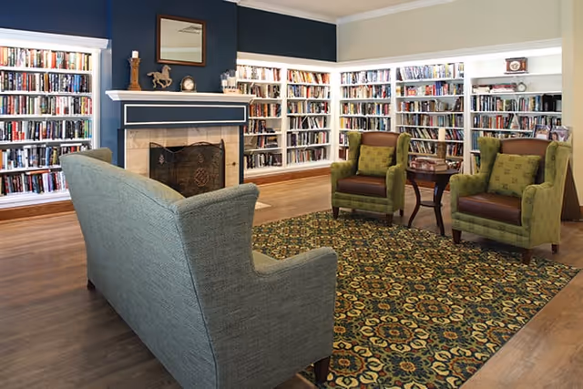 Cozy common room with bookshelves lining the walls, a fireplace, a sofa, two armchairs, and a patterned area rug.