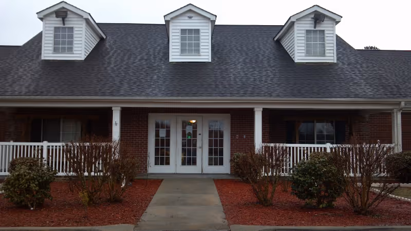 Front exterior view of a single-story brick building with a dark shingled roof and three dormer windows. The entrance features double glass doors with white framing, flanked by white railings and trimmed bushes on either side of a concrete walkway.