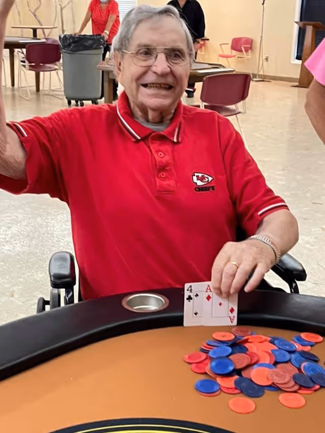 An elderly man in a red Kansas City Chiefs polo shirt sitting in a wheelchair at a poker table, smiling and holding playing cards (4 of clubs and ace of diamonds) with poker chips scattered on the table. In the background, there are chairs, a trash bin, and other people in a spacious room.