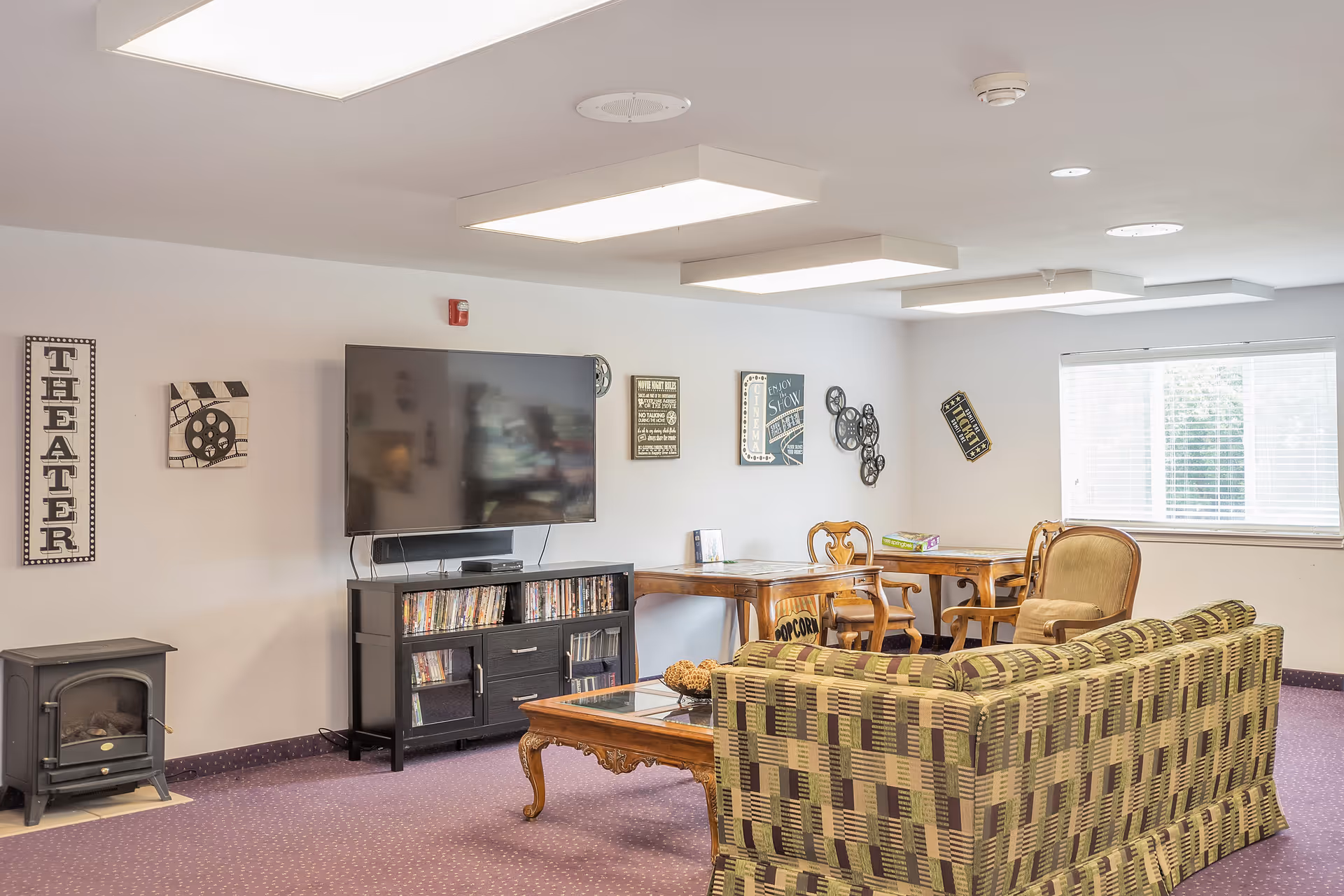 Well-lit common lounge with a wall-mounted TV, patterned sofa, armchairs, and a coffee table.