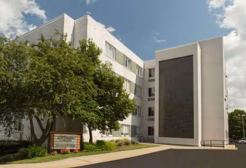 White multi-story senior living building with trees, a driveway, and a sign in front.