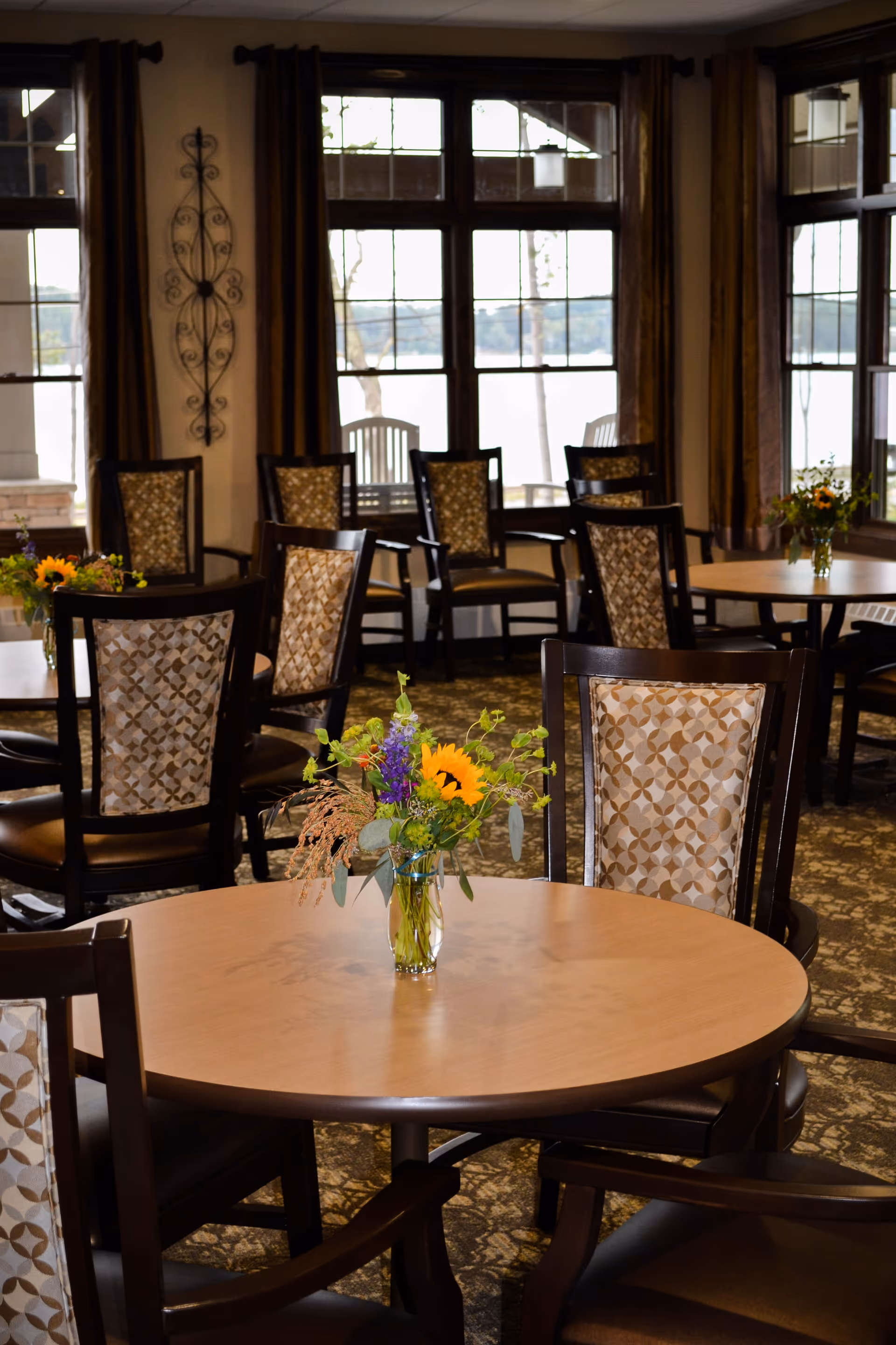 A cozy dining area with round wooden tables and cushioned chairs featuring patterned upholstery. Each table has a small vase with a colorful flower arrangement. Large windows with brown curtains allow natural light to fill the room and offer a view of trees and water outside.