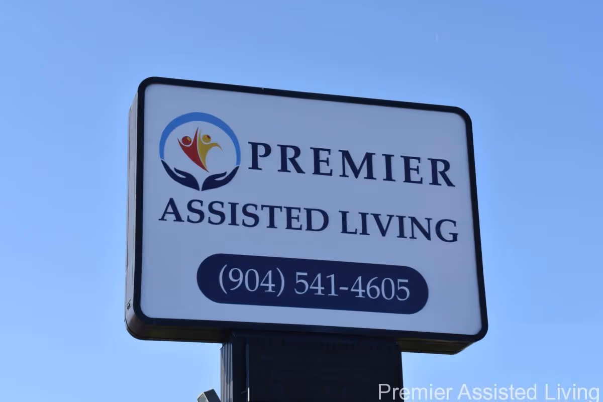 Outdoor sign for Premier Assisted Living with a logo featuring two stylized human figures in red and yellow above two hands, and a contact phone number (904) 541-4605, set against a clear blue sky.
