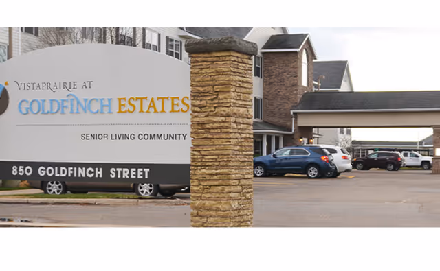 Entrance sign for Vista Prairie at Goldfinch Estates senior living community with a stone pillar, the building and parked cars visible behind it.