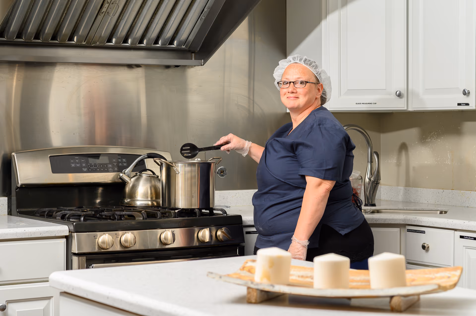 A woman wearing a hairnet, glasses, and gloves is cooking in a kitchen. She is stirring a large pot on a stainless steel stove with a kettle beside it. The kitchen has white cabinets, a countertop with a sink, and a decorative tray with three candles in the foreground.