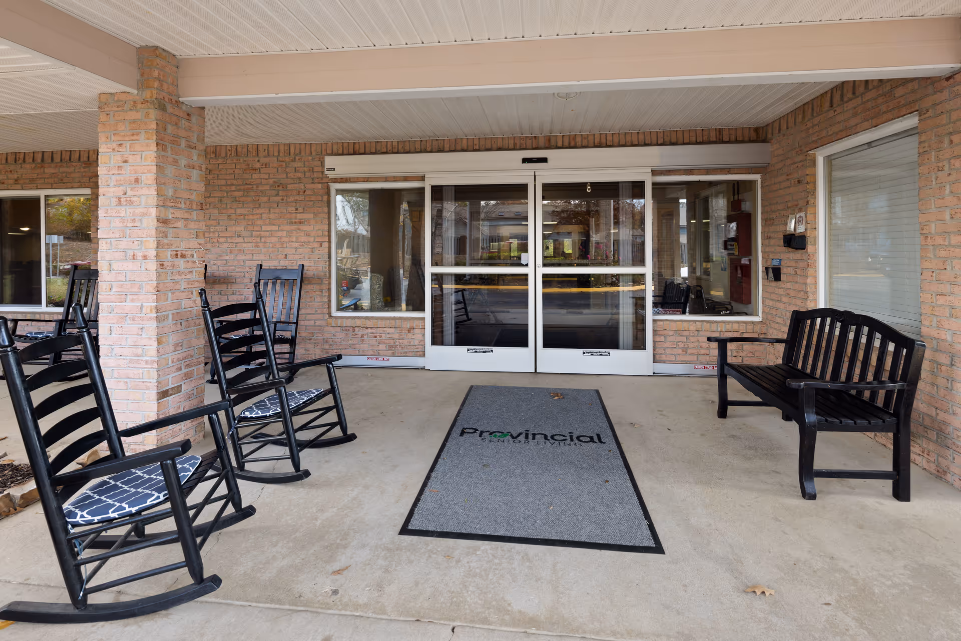 Covered entrance area of a senior living facility with brick walls, featuring black rocking chairs with cushions on the left and a black bench on the right. Automatic sliding glass doors lead inside, and a gray floor mat with the word 'Provincial' is placed in front of the doors.