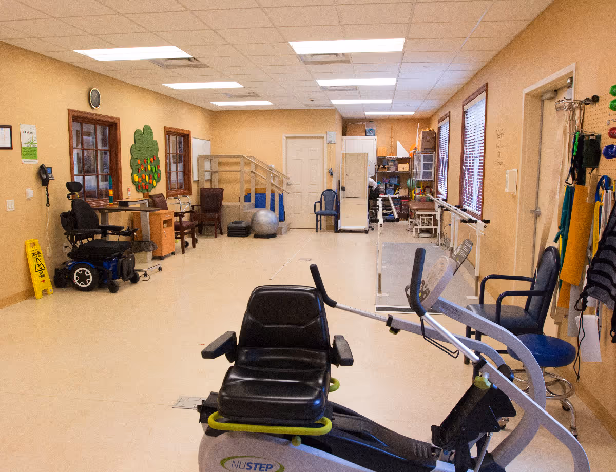 A rehabilitation or physical therapy room with exercise equipment including a NuStep recumbent cross trainer, a wheelchair, therapy chairs, parallel bars for walking exercises, a therapy ball, and various other therapy tools and equipment. The room has beige walls, a tiled ceiling with fluorescent lights, and windows with blinds.