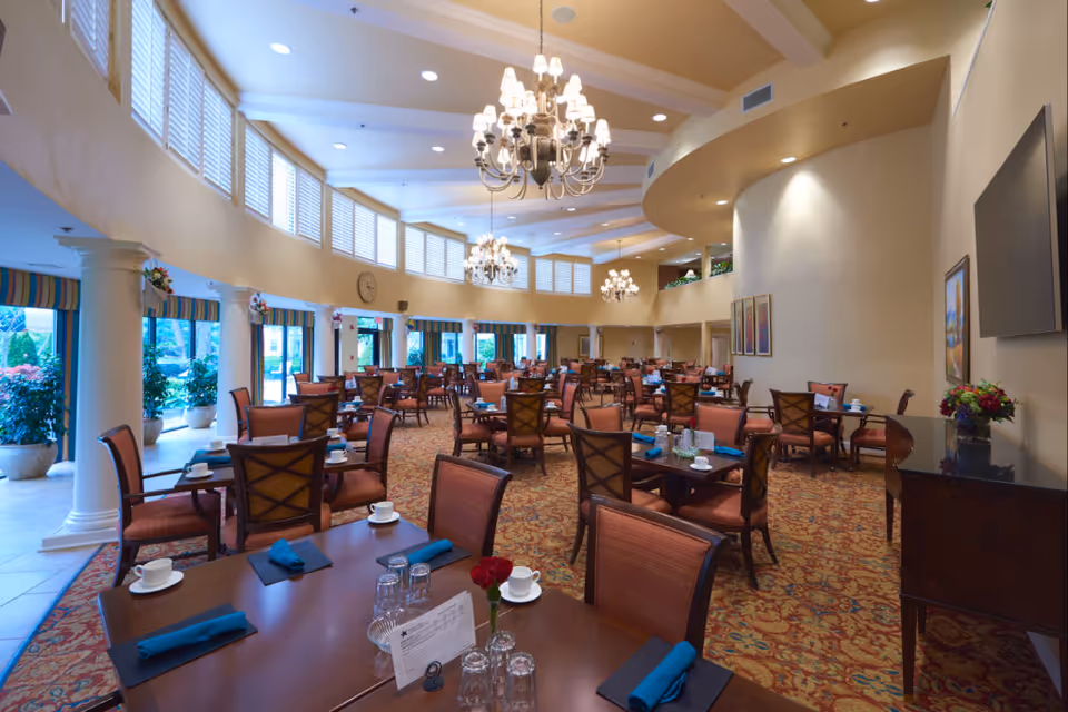 A spacious dining room in an assisted living facility with multiple tables and chairs arranged neatly. Each table is set with white cups, blue napkins, and glassware. The room features large windows with white shutters, elegant chandeliers hanging from the ceiling, and decorative columns along the side. There are plants visible outside the windows and framed artwork on the walls.