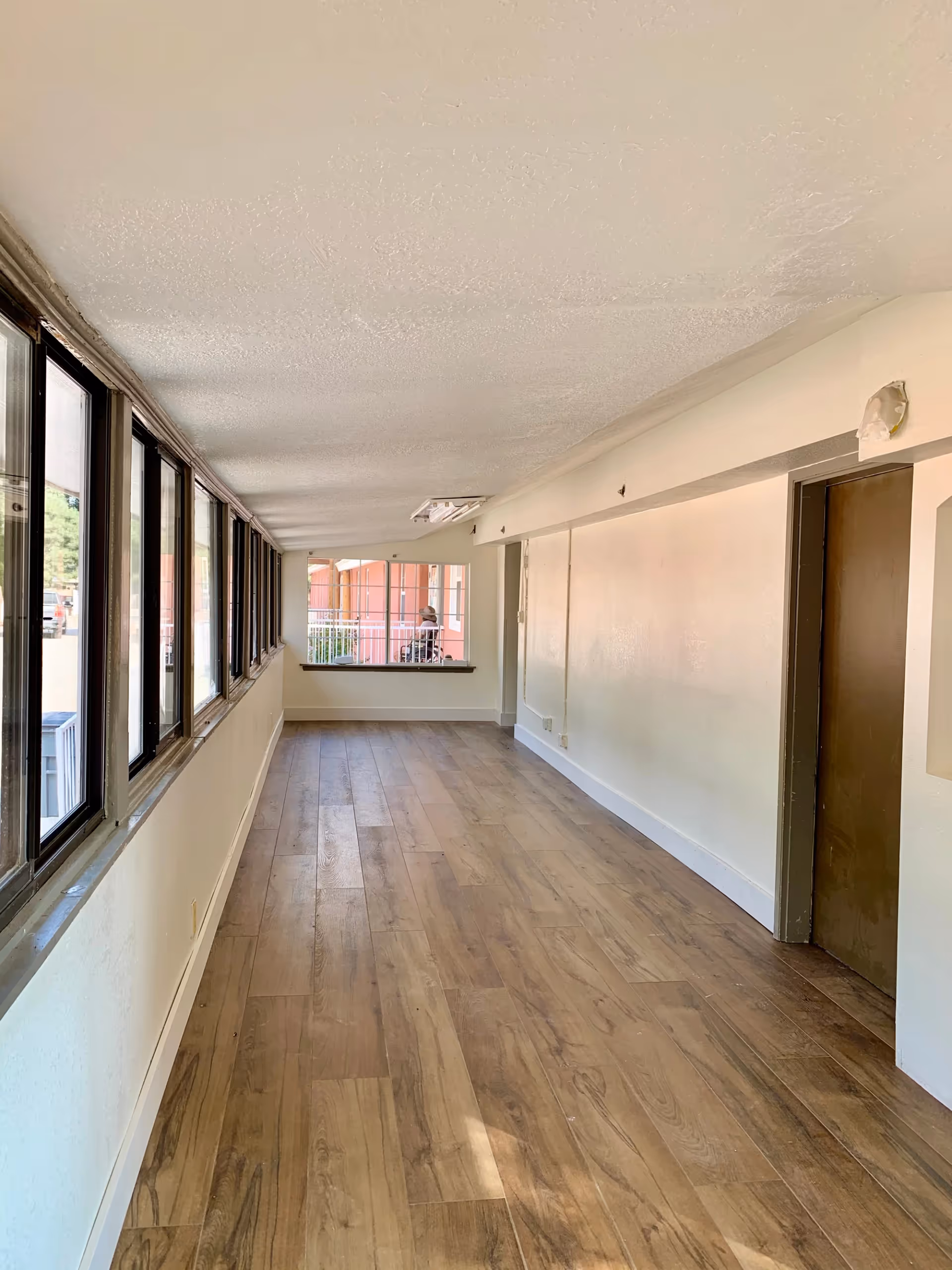 A long, narrow indoor hallway with wood-patterned flooring and large windows on the left side letting in natural light. The walls are painted white, and there is a brown door on the right side. At the end of the hallway, there is a window showing an outdoor area with a person sitting on a wheelchair.