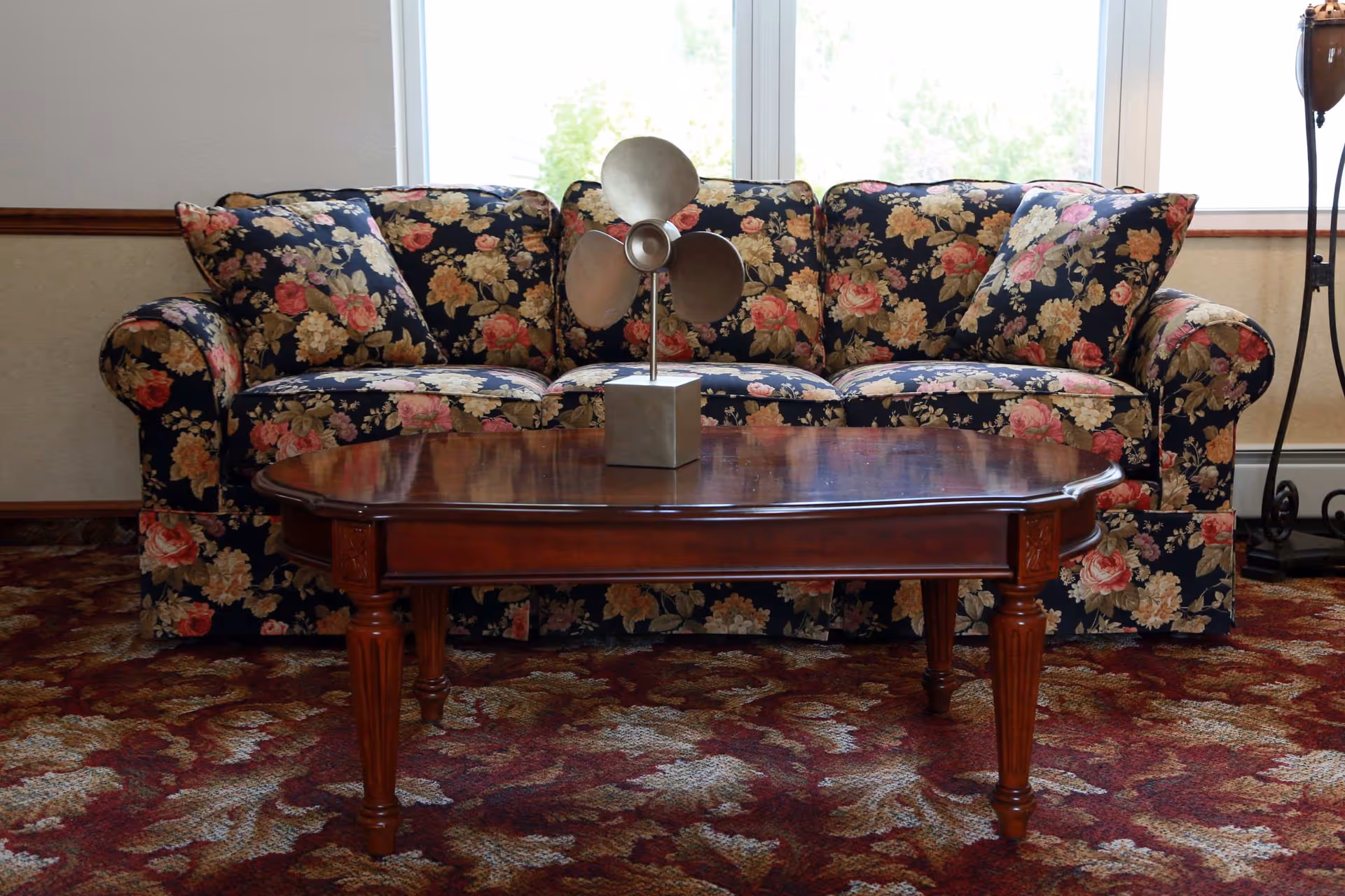 A floral-patterned sofa sits against a window with a wooden coffee table and a metal decorative sculpture on top, on a patterned carpet.