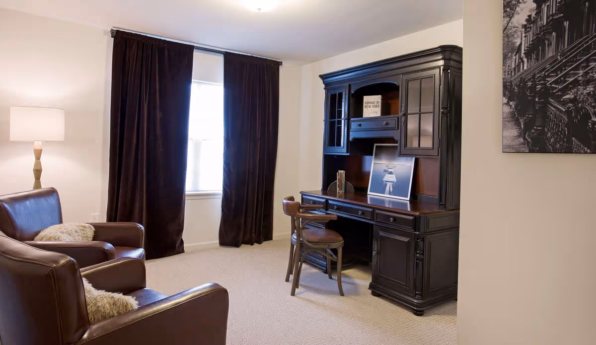 A cozy room with two brown leather armchairs with light-colored cushions, a tall floor lamp, a window with dark curtains, and a large dark wooden desk with a matching chair. The desk has decorative items and framed pictures on it, and a black and white photo hangs on the wall.