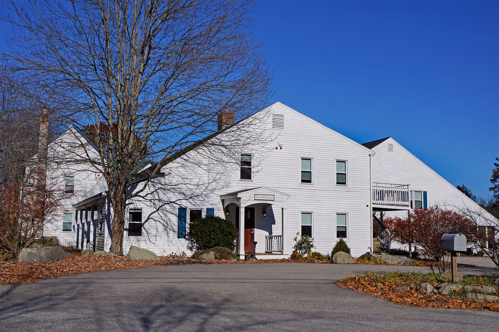 Exterior view of a white two-story building with multiple windows and a guest entrance. The building is surrounded by leafless trees and bushes, with a clear blue sky in the background. There is a paved driveway and a mailbox in the foreground.