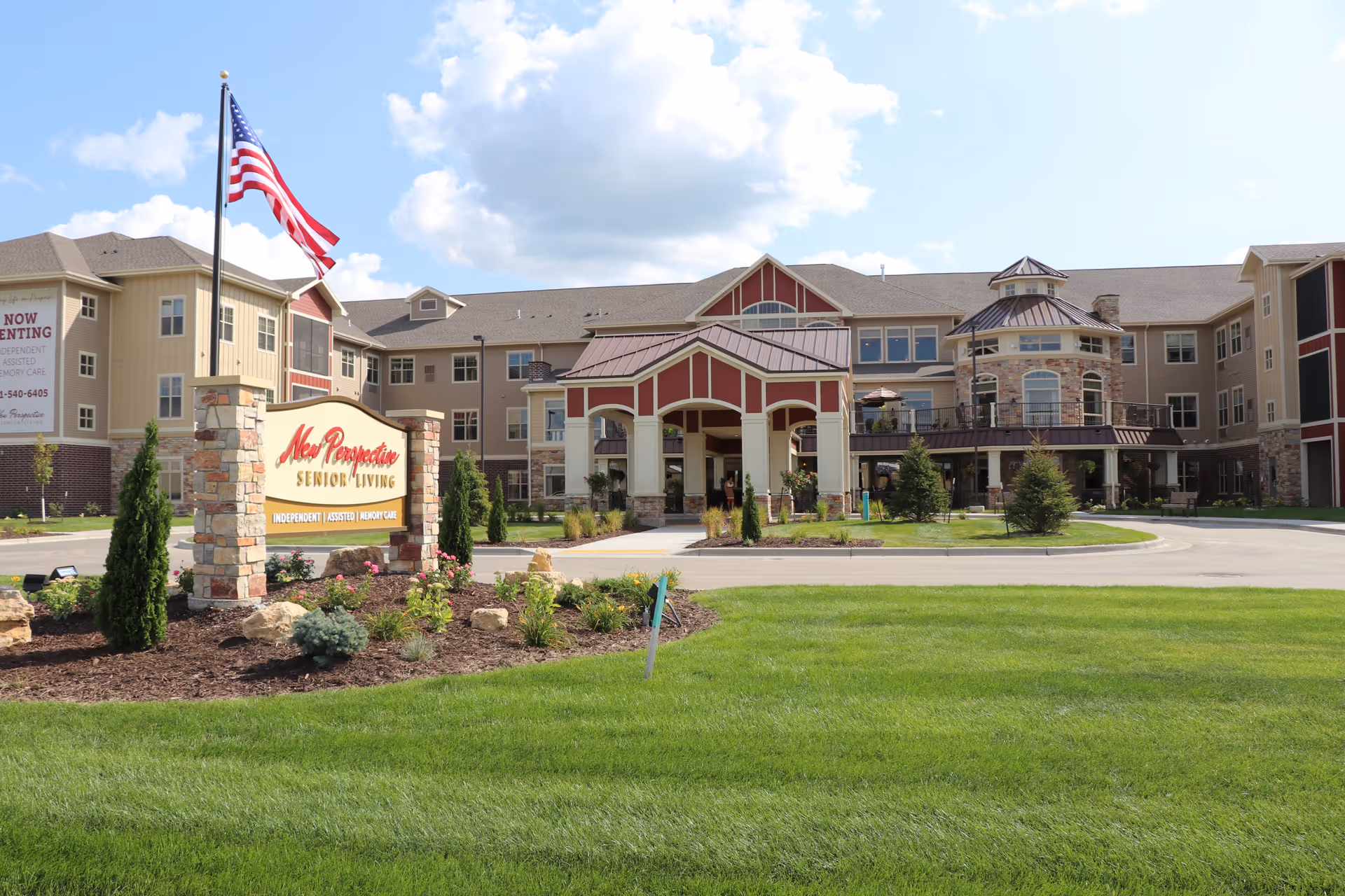 Exterior view of New Perspective Senior Living facility in West Fargo, showing a large building with beige and red accents, a covered entrance, landscaped garden with shrubs and flowers, an American flag on a flagpole, and a sign indicating independent, assisted, and memory care services.
