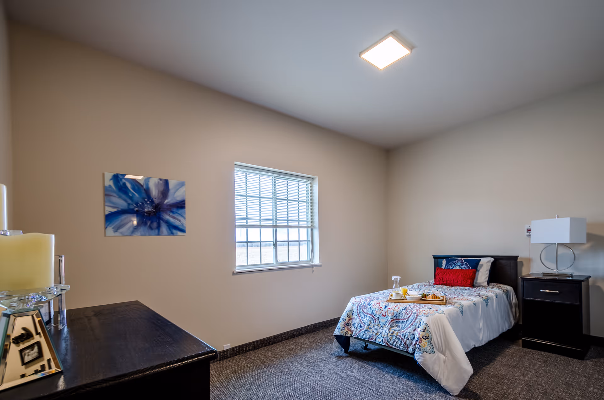 A simple, clean bedroom with a single bed covered in a white and blue patterned comforter, a red pillow, and a tray with breakfast items on the bed. There is a window with blinds on the beige wall, a dark wooden nightstand with a modern lamp, a dark wooden dresser with a candle, and a blue floral painting on the wall.
