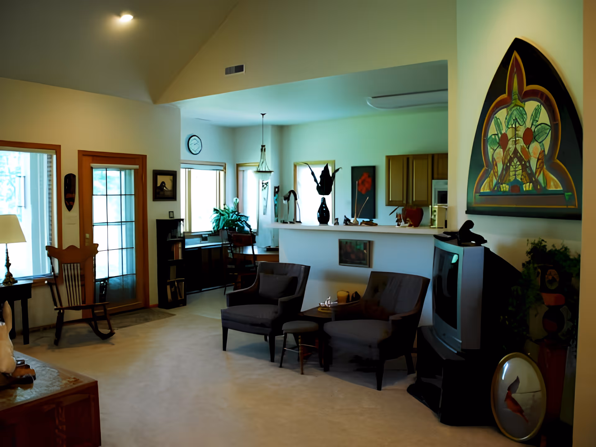 A cozy living room area with two gray armchairs and a small round table between them. There is a large TV on the right side, a colorful framed artwork above it, and various decorative items on the walls and surfaces. The room has beige carpeting, a wooden rocking chair near a window and door with glass panels, and a view into a kitchen area with wooden cabinets and a hanging light fixture.