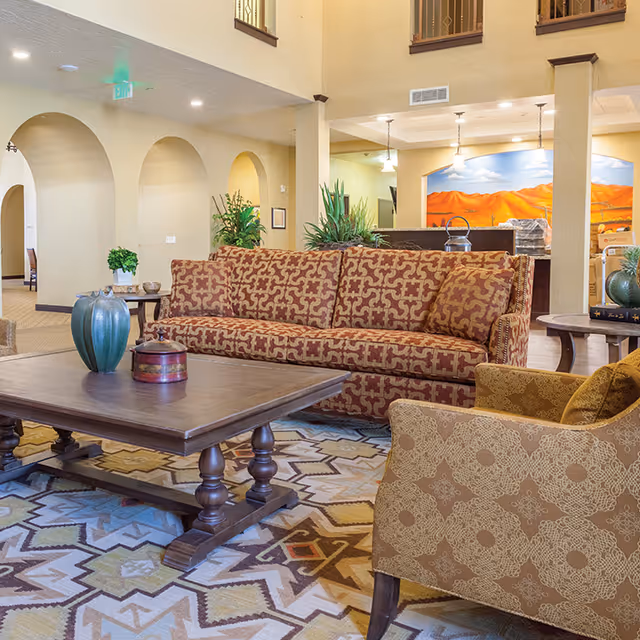 A cozy living room area in Jaidon Manor assisted living facility featuring a patterned sofa and armchair, a wooden coffee table with decorative items, potted plants, and a colorful patterned rug. The background shows arched doorways and a mural of a desert landscape with mountains.