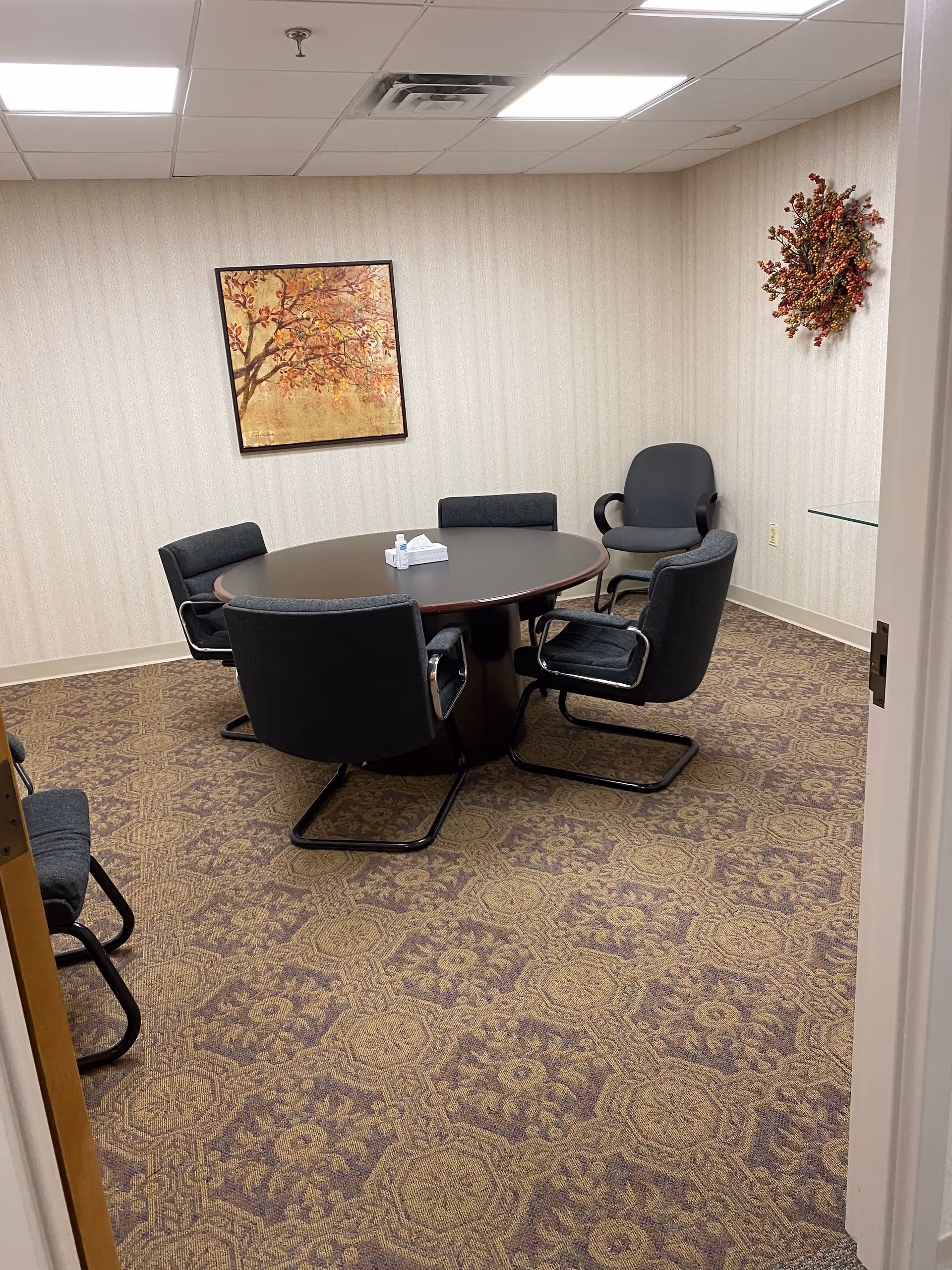 A small meeting or conference room with a round dark wood table surrounded by five black cushioned chairs. The room has patterned carpet flooring, light-colored wallpaper, a framed painting of a tree with autumn leaves on the wall, and a decorative wreath with red berries on another wall. A box of tissues is placed on the table.