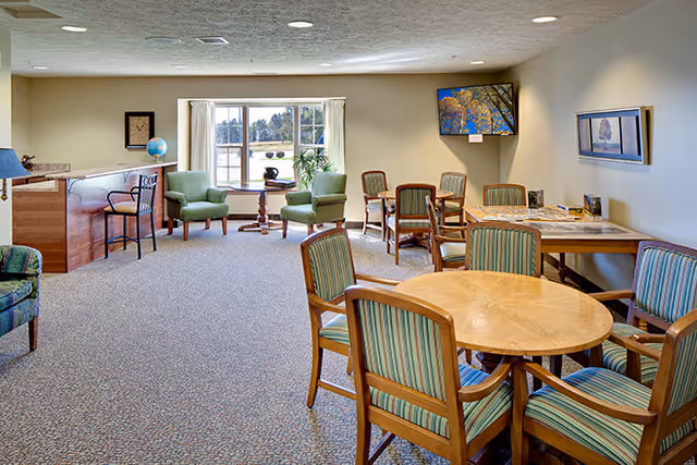 Bright common lounge in a senior living facility with round wooden tables and striped chairs, a seating area by a window, and a wall-mounted TV.