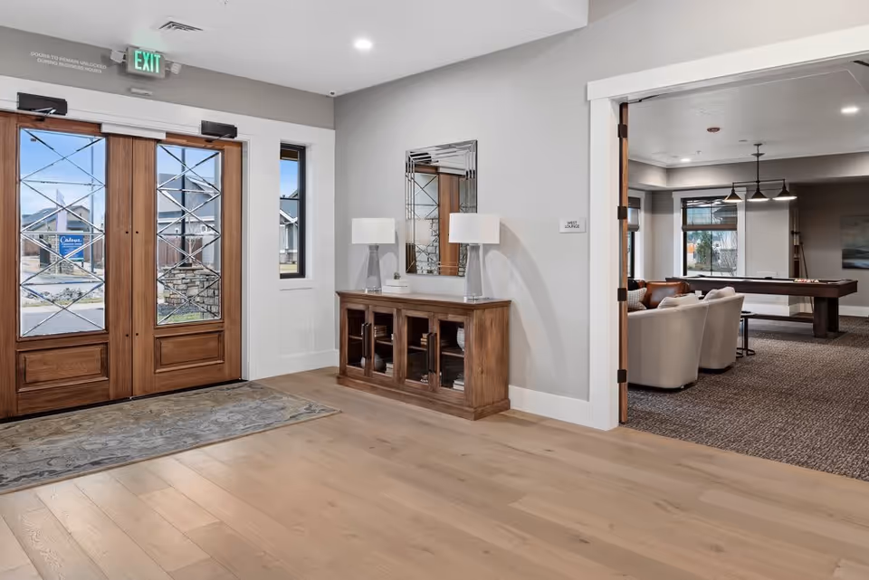 Entrance area of Cadence Independent Living featuring double wooden doors with glass panels, a wooden console table with two lamps and a mirror above it, and an open doorway leading to a lounge area with seating and a pool table.