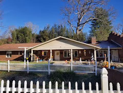 Single-story retirement center with a covered front porch, white picket fence, and tall trees.