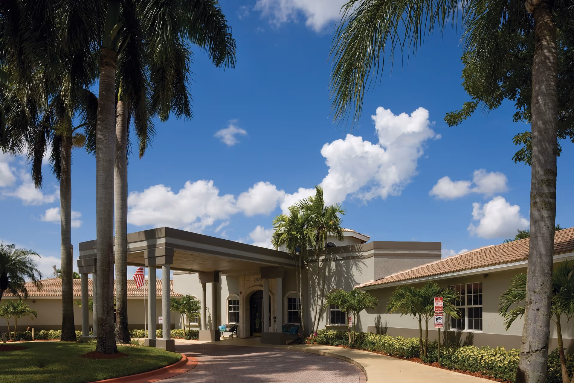Exterior view of HarborChase of Coral Springs facility entrance with a covered drop-off area, palm trees, and a clear blue sky with scattered clouds.