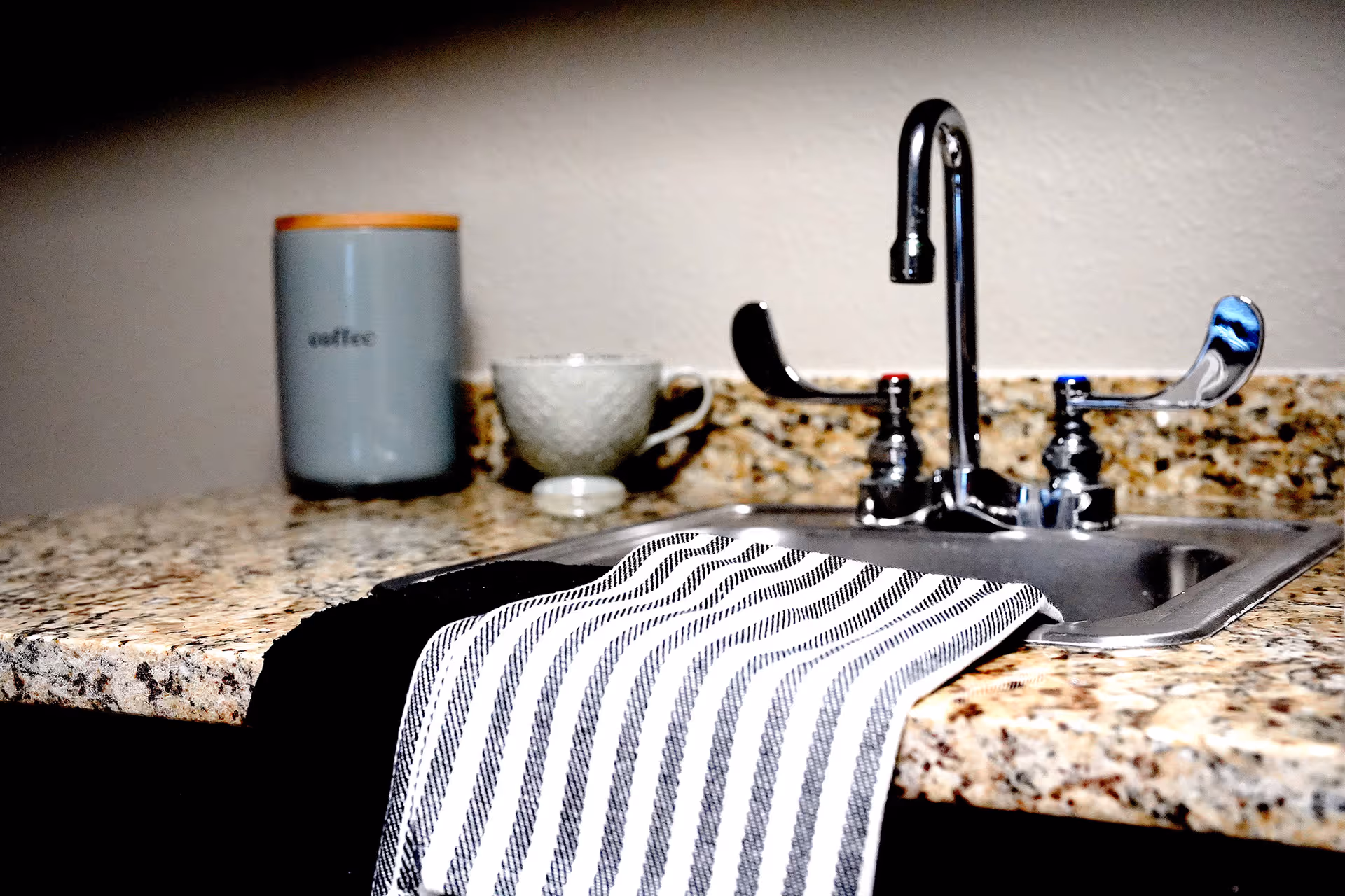 A kitchen sink with a chrome faucet and two lever handles, one with a red marker and the other with a blue marker. A black and white striped dish towel is draped over the edge of the sink. On the granite countertop behind the sink, there is a gray container labeled 'coffee' with an orange lid and a textured white coffee cup with a saucer.