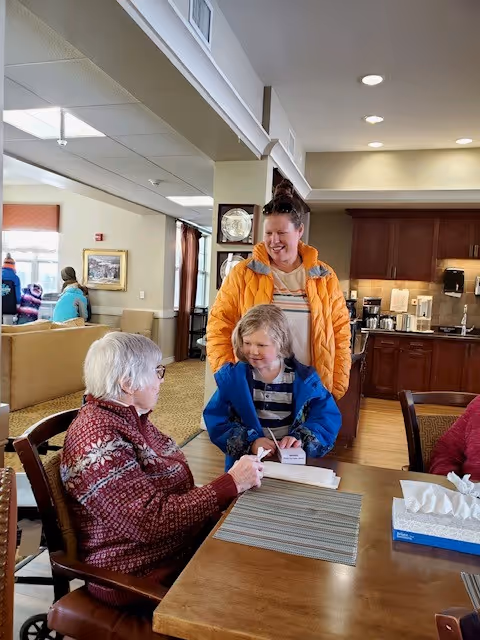 An elderly woman sitting at a wooden table in a common area, interacting with a young child and a woman standing next to the child. The elderly woman is wearing a red patterned sweater, the child is in a blue jacket, and the woman is in an orange jacket. The background shows a kitchen area with wooden cabinets and a coffee station, and other people sitting on couches in a well-lit room.