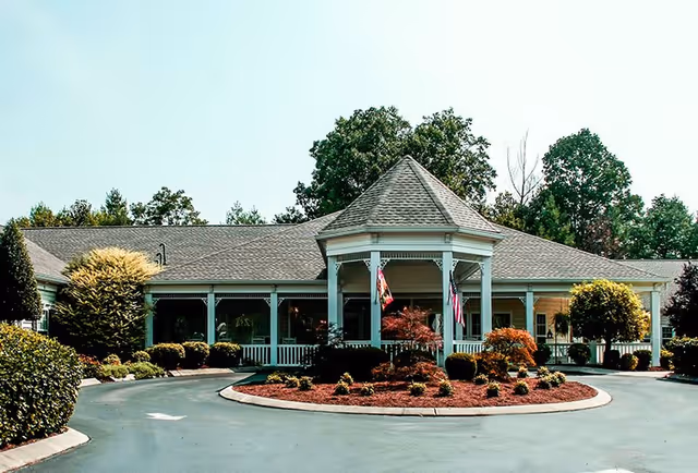 Front exterior view of Charter Senior Living of Cookeville building with a circular driveway, landscaped garden with shrubs and small trees, and a covered entrance with a peaked roof and white columns.