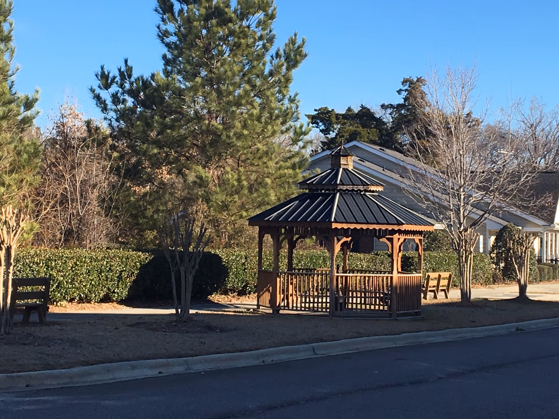 A wooden gazebo with a black metal roof situated on a grassy area near a paved road, surrounded by trees and bushes with a residential building in the background under a clear blue sky.
