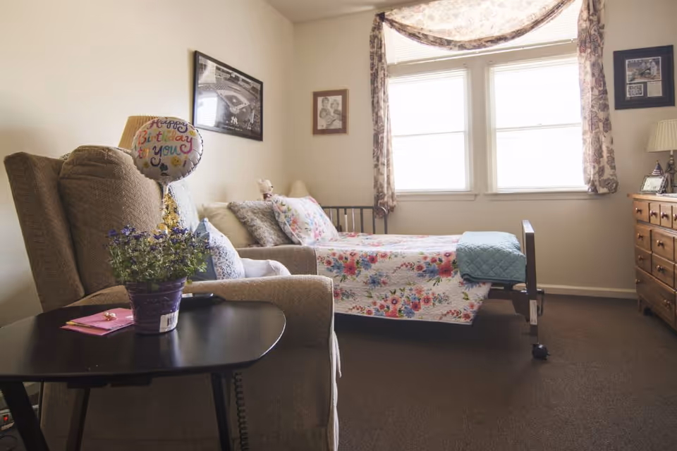 A cozy bedroom in a senior living facility with a single bed covered in a floral quilt, a cushioned armchair with a 'Happy Birthday to you' balloon and a potted plant on a small black table beside it. The room has two windows with floral curtains, a wooden dresser, and framed pictures on the walls.