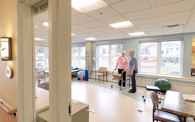 A bright room with large windows showing an elderly woman using a walker and a caregiver standing beside her. The room contains chairs, a table, and exercise equipment such as a large green exercise ball and a blue foam roller. The setting appears to be a rehabilitation or therapy area within a senior living facility.