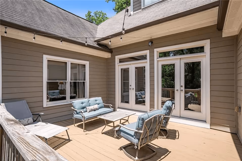 Outdoor patio area with cushioned seating including a loveseat, two chairs, and a lounge chair around a rectangular coffee table. The patio is surrounded by beige siding walls with two sets of white-framed French doors and a window. String lights hang above the seating area under a clear blue sky.